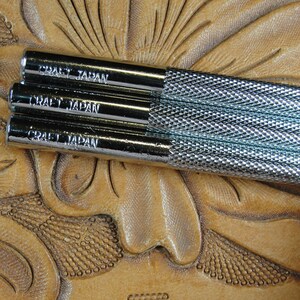 Puede incluir: Primer plano de tres herramientas de artesan&iacute;a en cuero met&aacute;licas con el texto "CRAFT JAPAN" en el lateral. Las herramientas tienen un agarre texturizado y est&aacute;n sobre una superficie de cuero marr&oacute;n con un dise&ntilde;o floral.