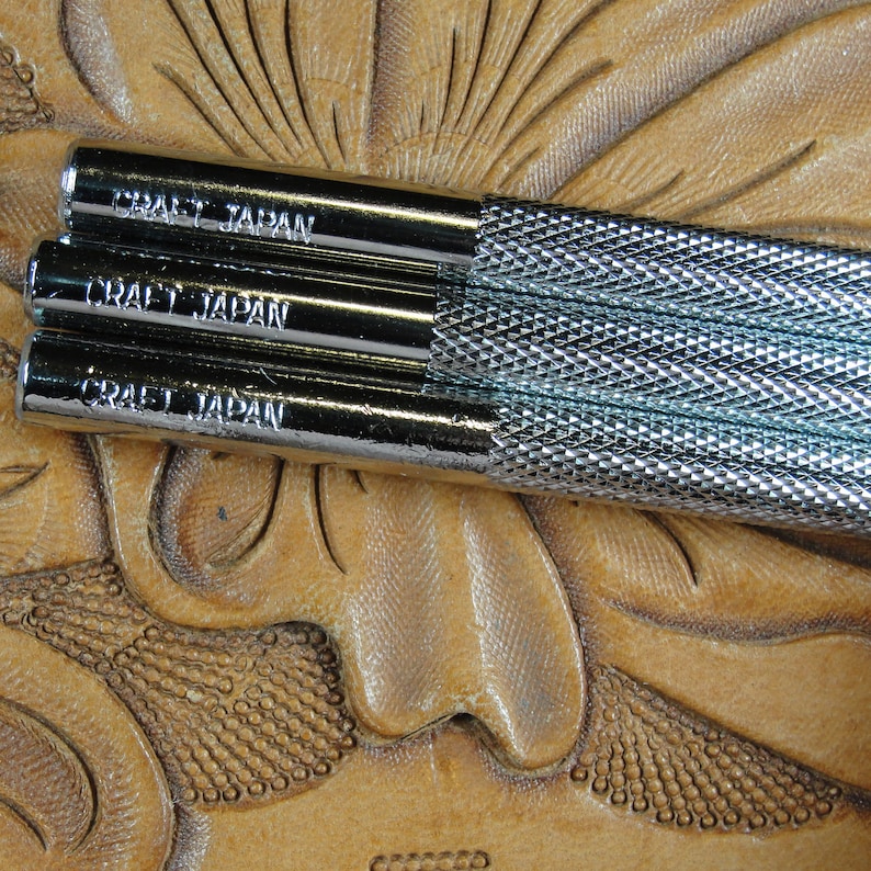 Puede incluir: Primer plano de tres herramientas de artesan&iacute;a en cuero met&aacute;licas con el texto "CRAFT JAPAN" en el lateral. Las herramientas tienen un agarre texturizado y est&aacute;n sobre una superficie de cuero marr&oacute;n con un dise&ntilde;o floral.