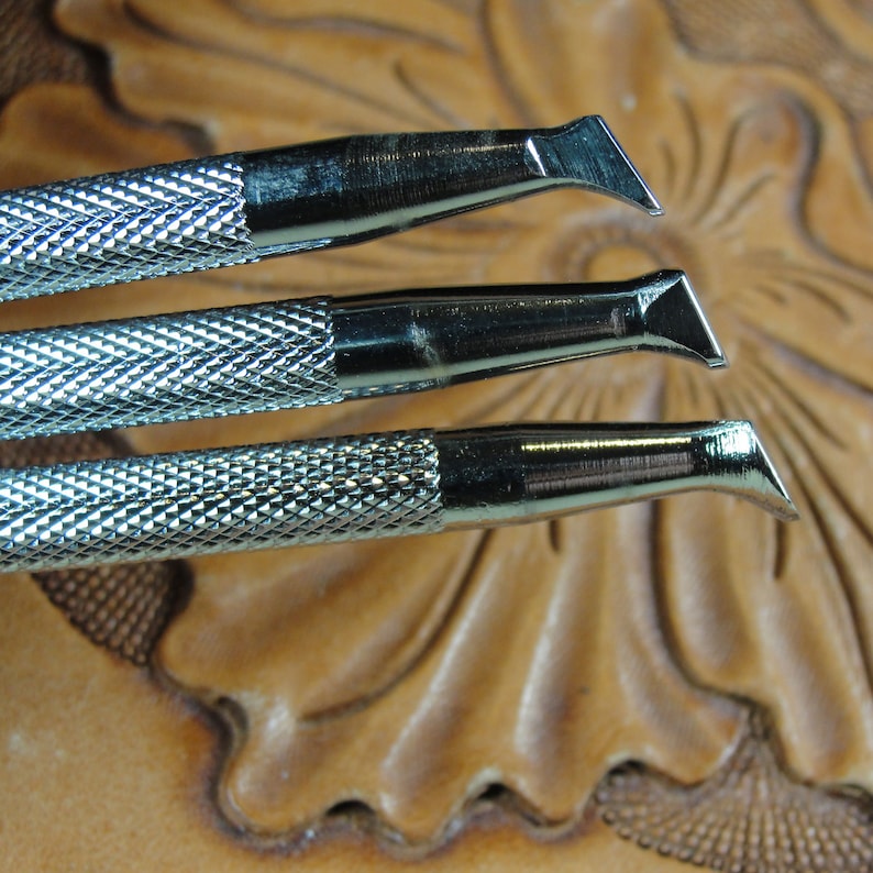Puede incluir: Primer plano de tres herramientas met&aacute;licas de marroquiner&iacute;a con cuchillas anguladas. Las herramientas tienen mangos texturizados con patr&oacute;n de diamante y est&aacute;n colocadas sobre un fondo de cuero tallado, lo que sugiere una artesan&iacute;a o pasatiempo.