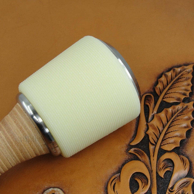 May include: A close-up of a leather working tool with a white, ribbed head and a wooden handle. The tool is resting on a piece of brown leather with a carved design.