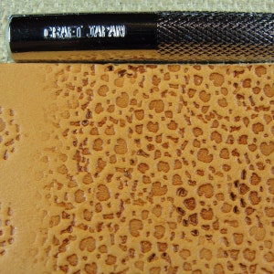Op de afbeelding: Close-up van een bruin lederen oppervlak met een textuurpatroon. Het oppervlak is ge&euml;mbost met een herhalend patroon van kleine cirkels. Een metalen gereedschap met de tekst "CRAFT JAPAN" is zichtbaar bovenaan de afbeelding.