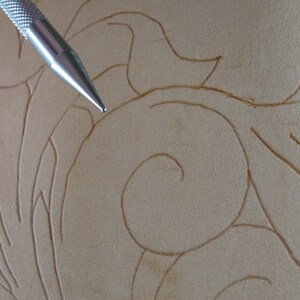 May include: Close-up of a metal tool used for leather carving. The tool is positioned over a piece of tan leather with a floral design. The leather has carved lines that create a swirling pattern.
