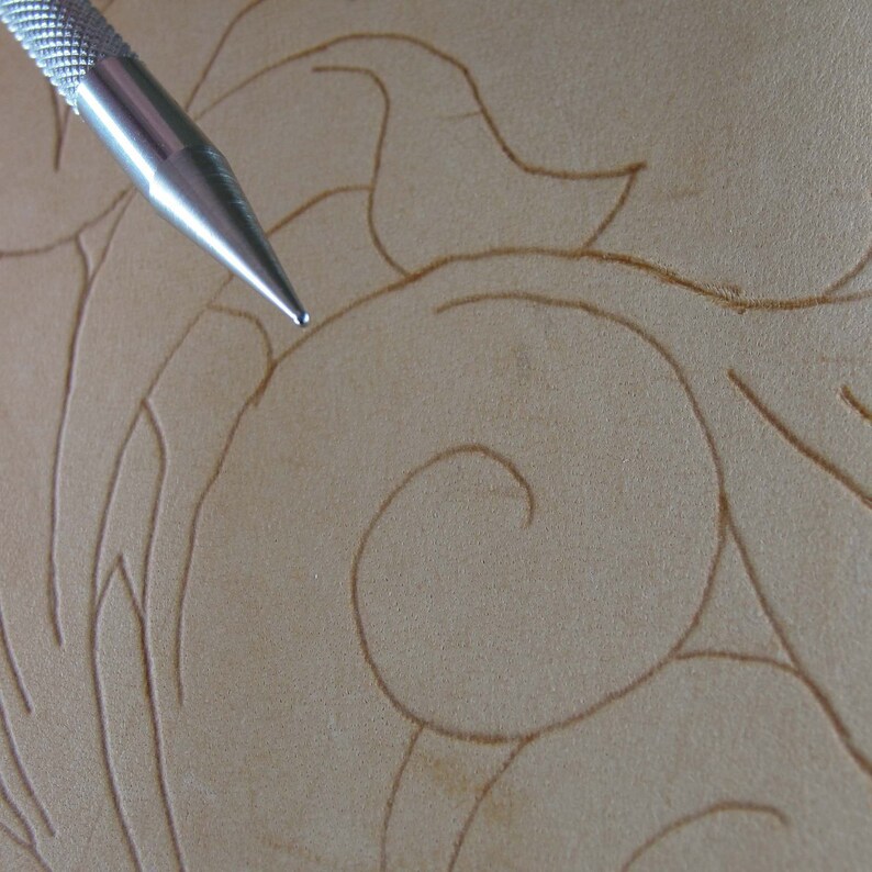 May include: Close-up of a metal tool used for leather carving. The tool is positioned over a piece of tan leather with a floral design. The leather has carved lines that create a swirling pattern.
