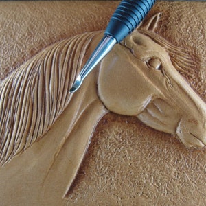 May include: A close-up of a leather carving of a horse's head with a tool used for leather carving resting on top of the carving.
