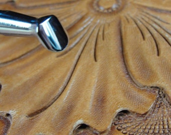 May include: Close-up of a leather carving with a floral design. A metal tool is positioned near the carved leather, which is tan with darker shading. The leather is embossed with a detailed flower and leaf pattern.