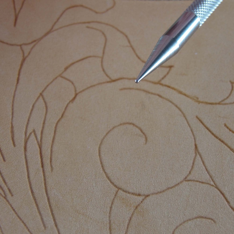 May include: Close-up of a leather crafting project with a metal tool. The tool is positioned over a light brown leather surface, with a detailed floral design drawn on it. The tool's tip is in focus, suggesting the process of tooling or carving.