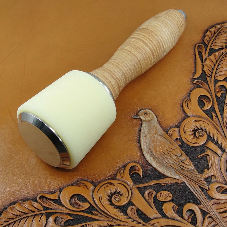 May include: A white leather mallet with a wooden handle and a silver metal band. The mallet is on a brown leather background with a carved design of a bird and floral patterns.