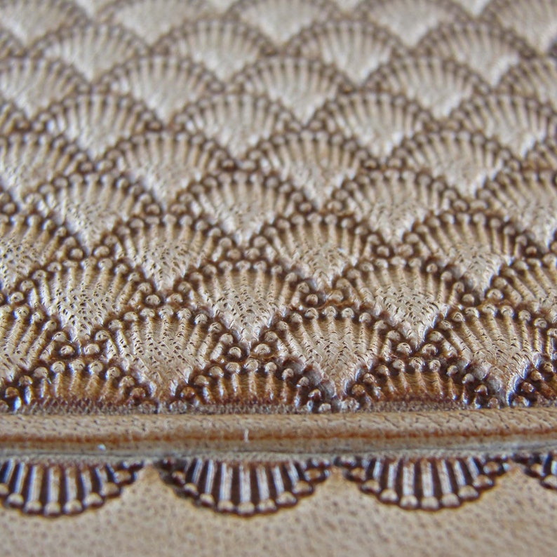 May include: Close-up of a brown leather surface with a textured, geometric pattern. The pattern is made up of small, raised triangles that create a chevron design. The leather has a light brown colour and a smooth, polished finish.