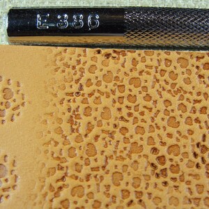 Op de afbeelding: Close-up van een bruin lederen oppervlak met een textuurpatroon van kleine, verhoogde cirkels. Het oppervlak is ge&euml;mbost met een patroon van kleine, verhoogde cirkels. Een metalen gereedschap met de tekst "E 386" is zichtbaar in de bovenkant van de afbeelding.