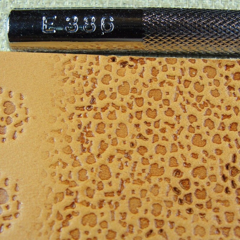 Op de afbeelding: Close-up van een bruin lederen oppervlak met een textuurpatroon van kleine, verhoogde cirkels. Het oppervlak is ge&euml;mbost met een patroon van kleine, verhoogde cirkels. Een metalen gereedschap met de tekst "E 386" is zichtbaar in de bovenkant van de afbeelding.