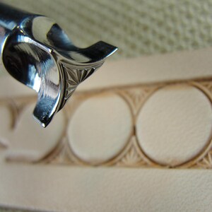 Puede incluir: Primer plano de una herramienta de marroquiner&iacute;a de metal brillante con un dise&ntilde;o de estrella. La herramienta est&aacute; colocada sobre un trozo de cuero de color claro con un patr&oacute;n circular tallado, mostrando artesan&iacute;a y detalle.