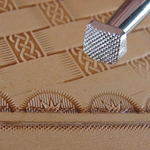 May include: Close-up of a leather stamping tool with a square head and a textured surface. The tool is being used to create a pattern on a piece of leather.