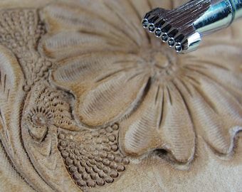 May include: Close-up of a leather carving tool with a row of small, pointed tips. The tool is positioned above a piece of leather that has been carved with a floral design.