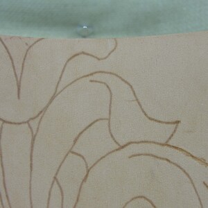 May include: Close-up of a beige leather surface with a floral design sketched in brown. The leather has a textured appearance. The top edge of the image shows a light green surface with a small, clear, round object.