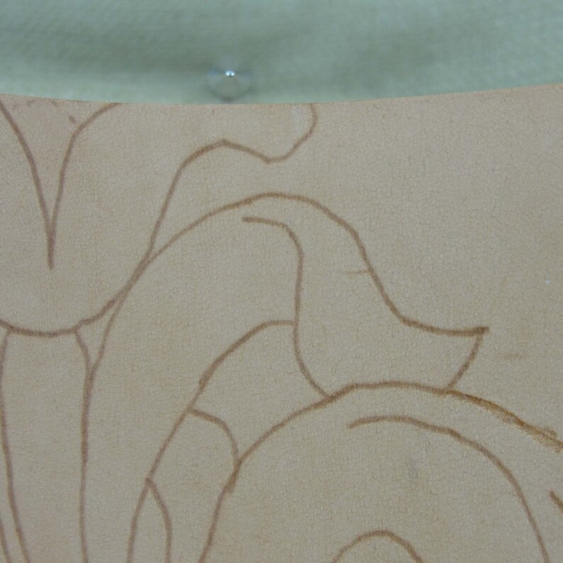 May include: Close-up of a beige leather surface with a floral design sketched in brown. The leather has a textured appearance. The top edge of the image shows a light green surface with a small, clear, round object.