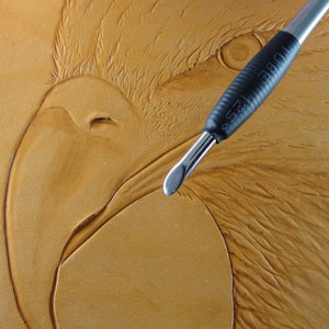 May include: A close-up of a leather carving tool being used to create a detailed image of an eagle's head on a piece of brown leather.