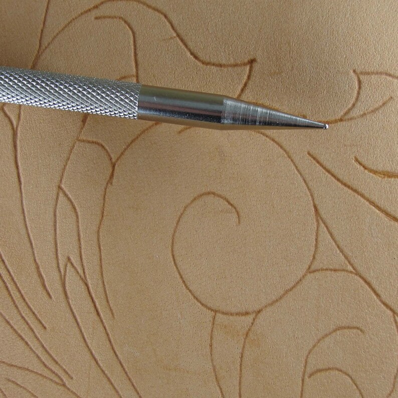 May include: Close-up of a silver metal swivel knife tool with a textured handle, positioned over a piece of tan leather. The leather has a carved floral design. The knife's sharp point is in focus, highlighting its use in leathercraft.