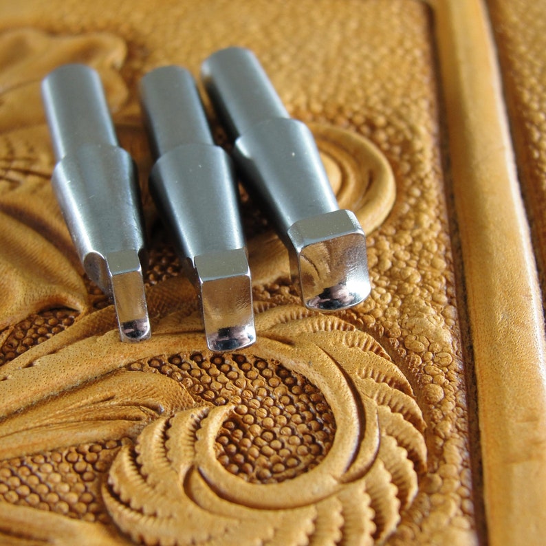 Puede incluir: Tres herramientas de estampado de cuero de metal plateado con bordes redondeados, dispuestas sobre un fondo de cuero marr&oacute;n con un patr&oacute;n decorativo.