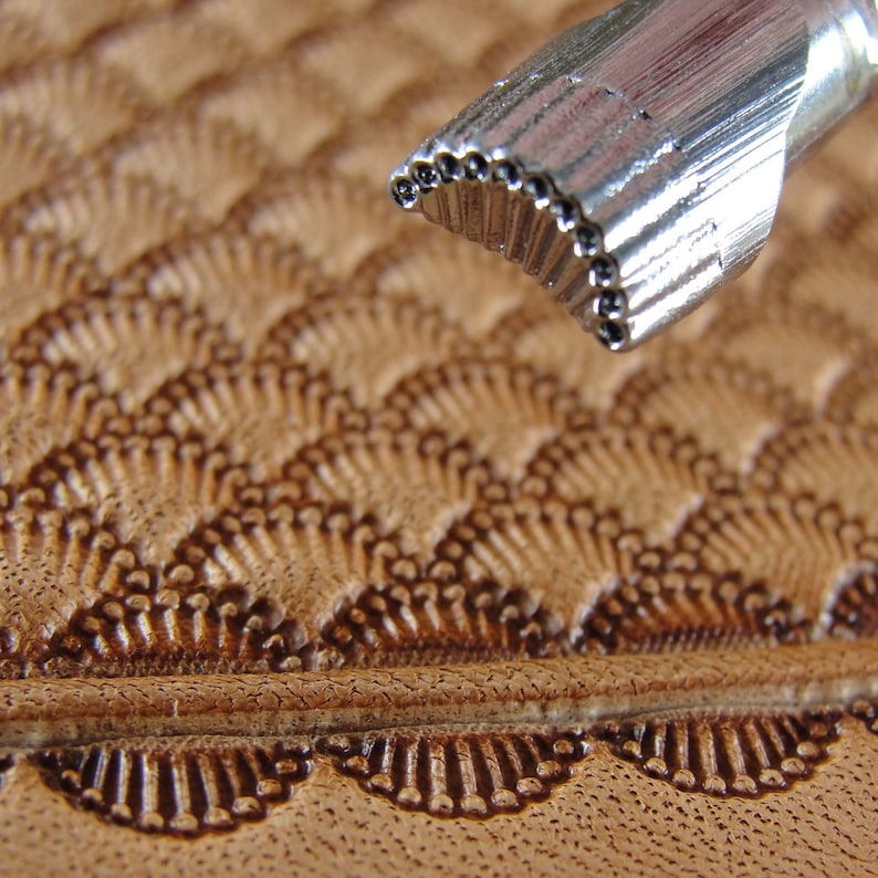 May include: Close-up of a metal leather stamping tool with a row of small, round punches. The tool is being used to create a decorative pattern on a piece of brown leather.