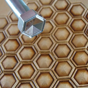Op de afbeelding: Een metalen leerstempelgereedschap met een geometrische zeshoekige kop is gepositioneerd boven een bruin leren oppervlak. Het leer is in reliëf voorzien van een herhalend honingraatpatroon, waardoor een textuureffect ontstaat. De tool wordt gebruikt voor leerbewerking.