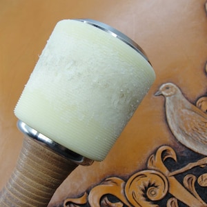 May include: A close-up of a leather working tool with a white rubber mallet head and a wooden handle. The mallet is resting on a piece of brown leather with a carved design.