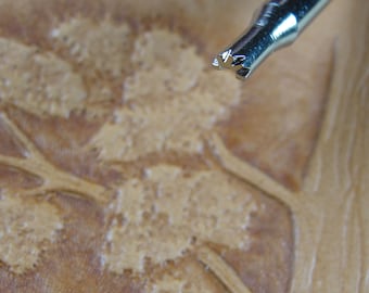 May include: Close-up of a metal leather stamping tool with a decorative tree design. The tool is held over a piece of tan leather with an embossed tree pattern. The tool's head has a detailed floral design.