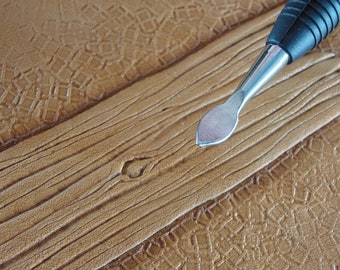 May include: A leatherworking tool with a black handle and a silver blade lies on tan leather. The leather has a wood grain texture, with a decorative geometric pattern in the background. The tool is for leathercrafting.