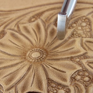 May include: A close-up of a leather carving tool with a textured, ridged tip. The tool is being used to create a floral design on a piece of leather.