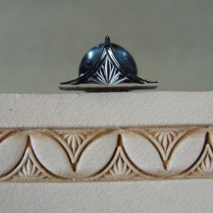 Puede incluir: Un objeto decorativo de metal plateado con un dise&ntilde;o triangular y una parte superior puntiaguda. El objeto se encuentra sobre una superficie clara y texturizada con un patr&oacute;n repetido de formas de hojas estilizadas.