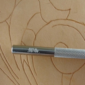 May include: Close-up of a silver metal leather working tool with a textured handle. The tool is marked "MADE IN JAPAN." The background is a tan leather surface with carved floral designs.