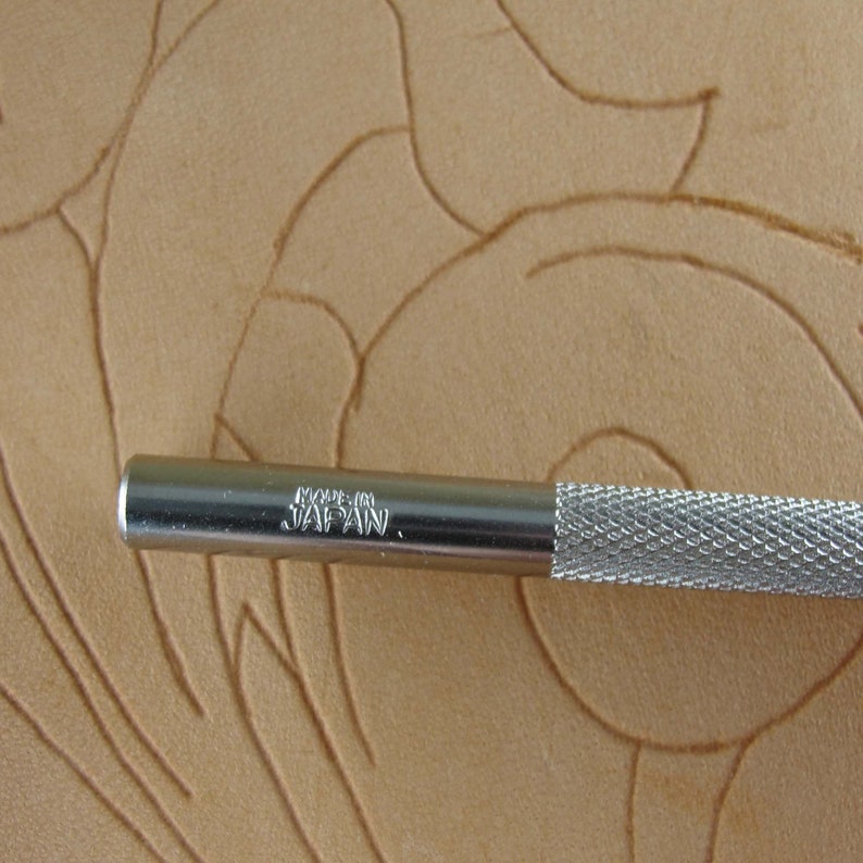 May include: Close-up of a silver metal leather working tool with a textured handle. The tool is marked "MADE IN JAPAN." The background is a tan leather surface with carved floral designs.