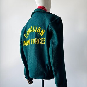 Könnte beinhalten: Dunkelgrüne Wolljacke mit gelbem Aufdruck "CANADIAN ARMED FORCES" auf dem Rücken. Die Jacke hat einen Knopfverschluss und einen rot-weiß gepunkteten Kragen.