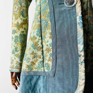 May include: A patterned jacket in green, blue, and gold, with a blue velvet panel and a large, clear button. The floral design is intricate, and the jacket has long sleeves. The overall style is vintage-inspired and elegant.