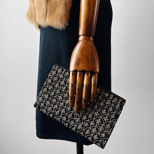 Puede incluir: Un clutch negro con un patrón floral dorado es sostenido por una mano de madera. El clutch es rectangular y tiene un diseño repetitivo. Un cuello de piel marrón es visible.