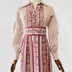 Peut inclure: Robe vintage avec un fond crème et un petit motif bordeaux répété. La robe présente des manches longues, un devant à volants et un imprimé floral bordeaux à la taille et sur la jupe. La jupe a des rayures verticales avec des motifs floraux.