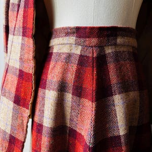 May include: A vintage skirt suit in a red, burgundy, and beige plaid pattern. The skirt has a fitted waist and a slight flare. The matching jacket is visible on the left, with a similar plaid design. The fabric appears to be tweed.