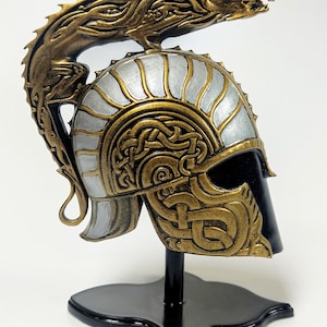 Peut inclure: Un casque doré et argenté avec un motif de dragon sur le dessus. Le casque est sur un support noir.