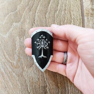 Può includere: Uno scudo di metallo nero con un bordo argentato e un albero bianco con radici e foglie. L'albero ha un anello attorno.