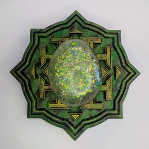 Peut inclure: Un mandala géométrique vert et or avec un cristal ovale transparent et iridescent au centre.