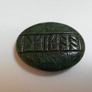 Op de afbeelding: Een groene, ovale steen met een gegraveerde inscriptie van het woord "TYKEN" in donkergroen.