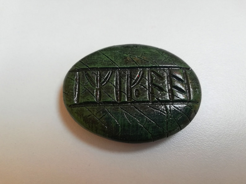 Op de afbeelding: Een groene, ovale steen met een gegraveerde inscriptie van het woord "TYKEN" in donkergroen.