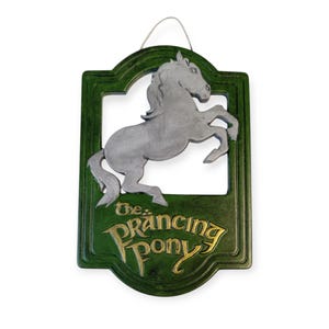 Puede incluir: Un letrero de madera verde con un caballo blanco saltando en el centro. El letrero dice "The Prancing Pony" en letras doradas.