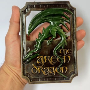 Peut inclure: Une sculpture de dragon vert avec une inscription dorée "The Green Dragon" sur une plaque en métal brun.