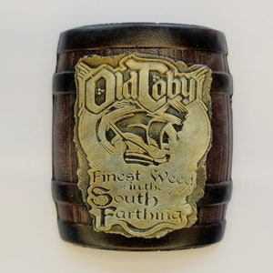 Może przedstawiać: Brązowa drewniana beczka z złotą etykietą, na której widnieje napis "Old Toby Finest Weed in the South Farthing".