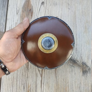 Op de afbeelding: Een bruin leren schild met een metalen rand en een gouden en zwarte centrale boss. Het schild wordt in een hand gehouden.