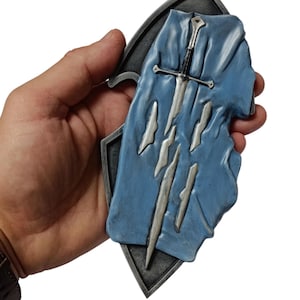 May include: A decorative metal shield featuring a blue fabric overlay and a silver sword. The shield has a dark grey border and a triangular base. The blue fabric has several silver slashes.