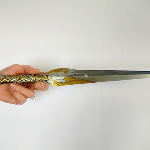 May include: An ornate dagger with a silver blade and a gold and silver handle. The handle features intricate braided and floral designs. The dagger is held against a white background.
