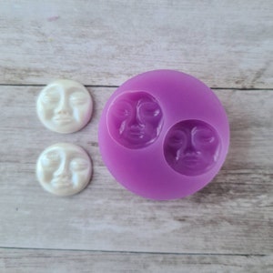 May include: A purple silicone mold with two moon face impressions, next to two white moon face cabochons. The moon faces have closed eyes and detailed facial features. The cabochons have a pearlescent finish.