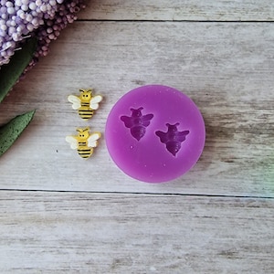 May include: A close-up of a purple silicone mold with two bee-shaped cavities, next to two yellow and black bee-shaped embellishments. The bees have white wings and black stripes. The background is a light wood grain.
