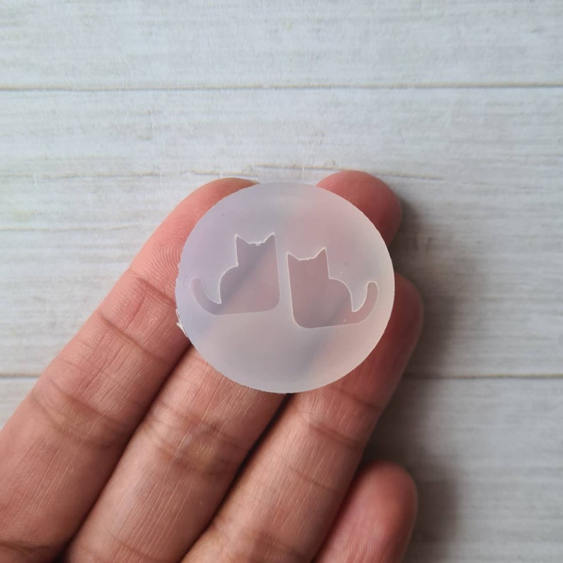 May include: A clear, circular silicone mold featuring two cat silhouettes side-by-side. The mold is held in a hand, with a light-colored wooden surface in the background. The cat shapes are detailed, with visible ears and tails, perfect for crafting.